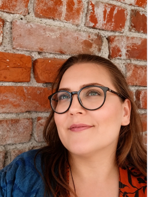 Nainen nojaa tiiliseinää vasten, käyttää mustia pyöreitä silmälaseja ja katsoo kameraan. kehopositiivisuus, Hiidetär